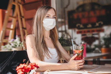 Portrait of a blonde student girl sitting alone in a cafe wearing a medical mask, holding a glass of drink in her hands, quarantine, loneliness during quarantine