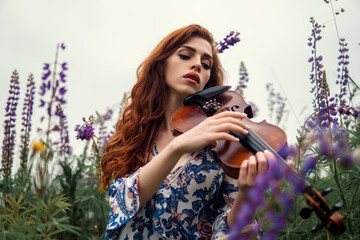Beautiful romantic girl with red hair and blue dress holding violin on nature field of  flowers....