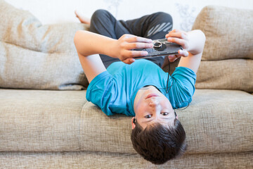 Attractive youngster guy wearing blue t-shirt lying on his back on couch and holding smartphone in hands, browsing social media