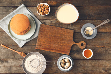 Tasty pancakes with ingredients and board on wooden background