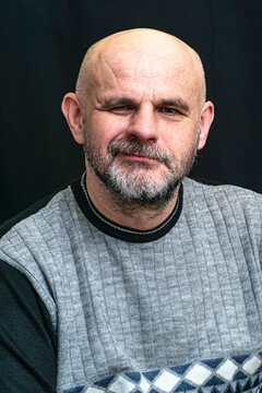 middle-aged bald bearded man grins contemptuously against the dark background