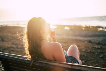 Happy beautiful girl viewed from back sit down on a bench and enjoy the sunset at the beach -...