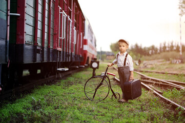 Sad boy, dressed in retro style, stands at the train station with suitcase and bike.