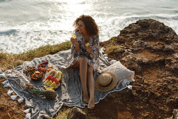 Happy curly hair woman sit on blanket on sea cliff, hold slices of lemon, enjoy picnic near the sea. Summer concept