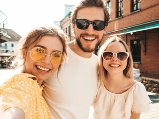 Group of young three stylish friends in the street.Man and two cute girls dressed in casual summer clothes.Smiling models having fun in sunglasses.Women and guy making photo selfie on smartphone