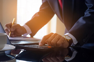 Businessman in suit, hand using calculator, analysing business data paper and work on laptop computer with cup of coffee on the table at modern home office.