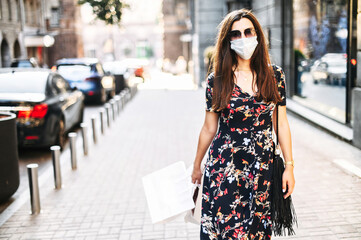 Shopping with protective measures after quarantine is ended. Woman walks on the city street with a medical mask on the face, paper shopping bags in her hands