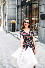 Obraz premium Vertical portrait of happy beautiful girl, she is walking on the street with a shopping bags and looks at camera cheerfully