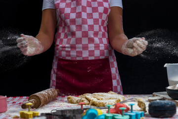 Girl claps hands and spraying flour over dough on dark background. Professional girl baker in apron is preparing cookies
