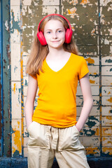 Youth and Teens Lifestyle. Positive Teenage Girl In Red Wireless Headphones Posing in Casual outfit Against Grunge Wall In Summer Park Outdoor.