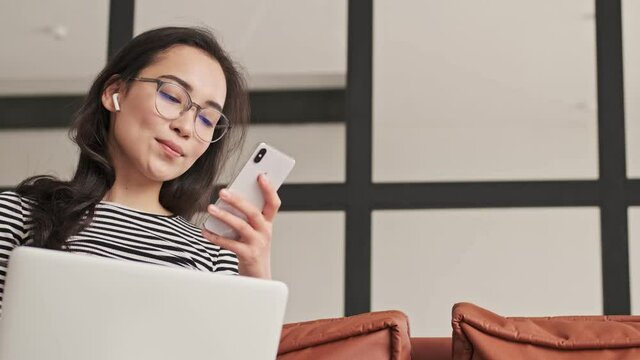 Smiling pretty asian woman in eyeglasses using smartphone while sitting with laptop computer on sofa at home