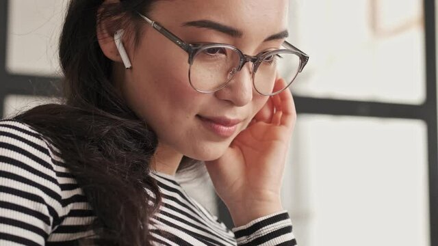 Close up view of Smiling pretty asian woman in eyeglasses and earphones sitting on sofa at home