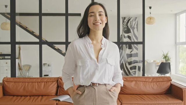 Cheerful elegant pretty asian woman posing with arms in pockets and looking at the camera at home
