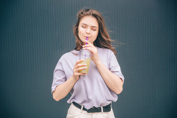 Young stylish trendy woman isolated over grey blue background. Dreamy girl drink cocktail or lemonade and enjoy iw with closed eyes. Stand alone and poses. Tasty delicious drink.