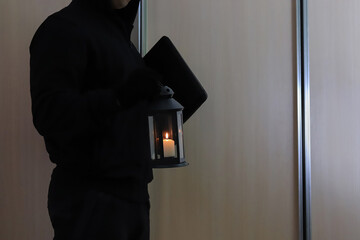 A thief in a black hood and gloves with a lamp with candles in someone else's dark apartment. Copy...