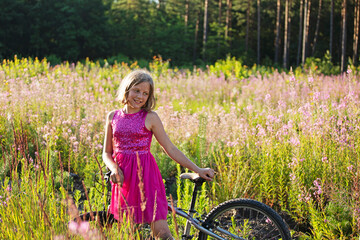  happy girl in a purple dress stands in a field of fireweed in a meadow, leaning on a bicycle on a summer evening day. self-isolation.