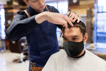 grooming, hairdressing and health safety concept - man wearing face protective medical mask for protection from virus disease and barber with comb and scissors cutting hair at barbershop