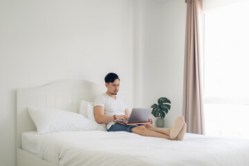 Asian man lying on bed work on his laptop in cozy white bedroom.