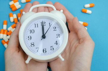 Women's hands hold a white alarm clock on a background of scattered tablets, capsules on a blue background. concept of time, health.