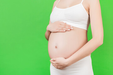 Cropped image of pregnant woman's hands embracing her abdomen at green background. Future mom is wearing white underwear. Expecting of a baby. Copy space