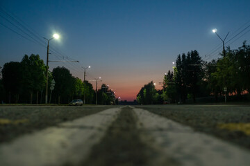 Fototapeta na wymiar Paved road on the outskirts of the city at sunset, on the sides of green trees grow, lights are burning