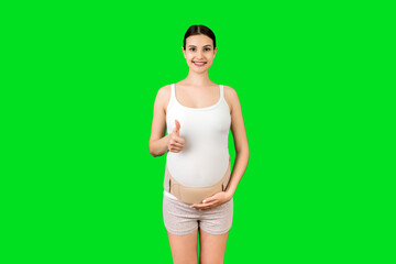 Portrait of young pregnant woman wearing bandage on her belly and showing thumb up sign at green background with copy space. Orthopedic abdominal support belt concept