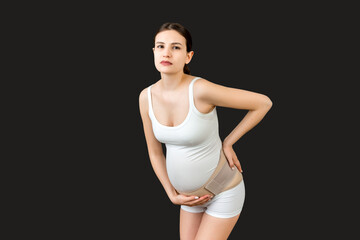 Obraz premium Portrait of pregnancy belt dressed on suffering pregnant woman for reducing pain in the back at black background with copy space. Orthopedic abdominal support belt concept