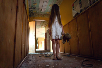 girl in a white dress with long hair and a Teddy bear in her hands stands in the middle of a creepy corridor in an abandoned building. Concept of horror, mysticism