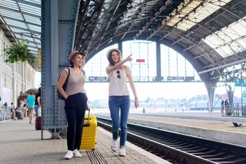 Happy mother and teenager daughter walking with suitcase at railway station