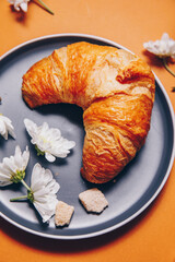 croissant with small white chrysanthemum flowers on an orange background. breakfast with a croissant on a gray plate