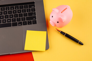 Moneybox near laptop and yellow sticky note, place for text. Finance and budget concept. Piggy bank in pink color with gadgets and stationery on colorful background