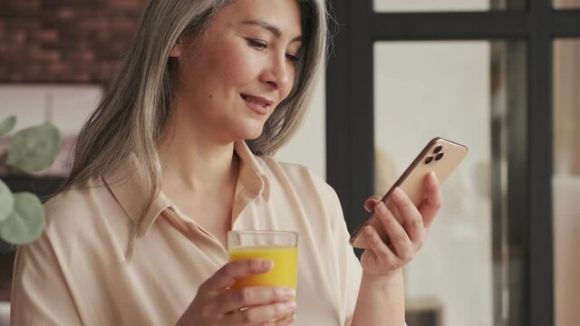 A happy pleased mature woman is drinking orange juice while using her smartphone in the kitchen at home