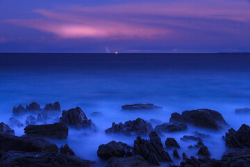 Deep blue ocean under a purple dusk sky. A distant flash of lightning is visible on the horizon.  Long exposure