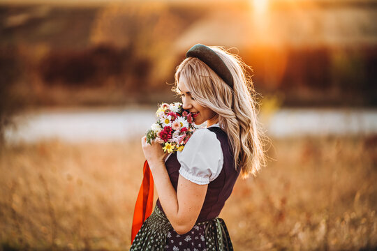 Pretty blonde girl in dirndl, standing outdoors in the field, holding bouquet of a field flowers.