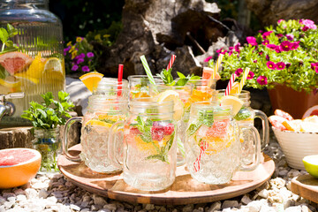 Infused water with lemon, grapefruit, lime and mint in glass cocktail mugs stands on a rotating wooden tray in the open air. Sunny day, bright pink and raspberry flowers in the background, relaxation.
