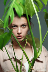 Fototapeta premium A girl in a pink jacket in black jeans holds in her hands a monstera flower in a pot posing on a gray background.
