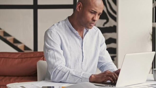 A good-looking african american man is using his laptop during working at home sitting at table in living room at home