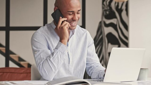 A happy pleased african american man is talking on his smartphone while working with laptop computer sitting at table at home