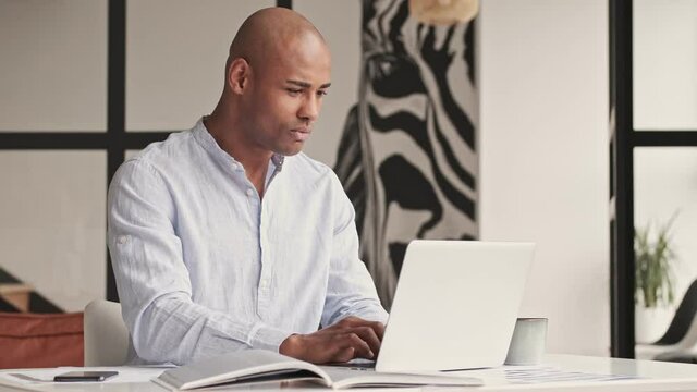 A confident focused african american man is working with laptop computer and smartphone sitting at table at home