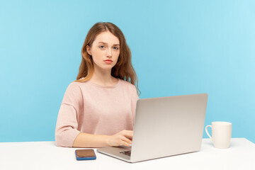 Naklejka premium Smart serious young pretty businesswoman in casual clothes looking at camera with responsible confident expression, working on laptop at home office. indoor studio shot isolated on blue background