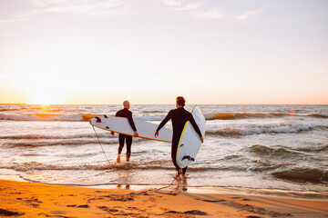 Two young male surfers in black wetsuits with longboards going to water at sunset ocean. Water...