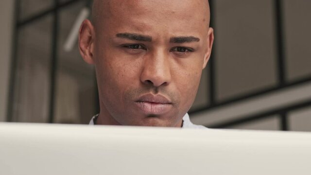 A close-up view of a concentrated african american man is using his silver laptop sitting at table at home