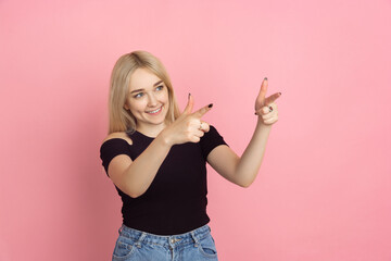 Pointing at side. Portrait of young caucasian woman with bright emotions on coral pink studio background. Blonde female model. Concept of human emotions, facial expression, sales, advertising, youth.