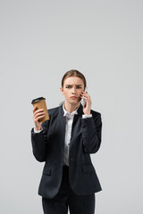 confused young businesswoman with coffee to go talking on smartphone isolated on grey