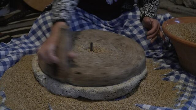 The ancient quern stone hand mill with grain. The woman grinds the grain into flour with the help of a millstone. Men's hands rotates a millstone. Old grinding stones turned by human hands
