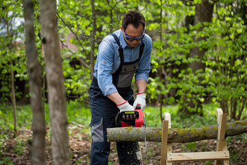 Close up view of woodcutter lumberman in working uniform sawing small tree trunk on sawhorse with...