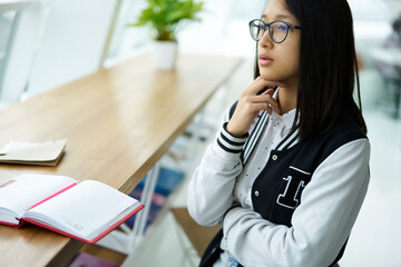 Pondering pretty asian girl with eye glasses dressed in jacket studying in library and thinking about new ideas for school project.Thoughtful chinese student sitting in coffee shop with notebook