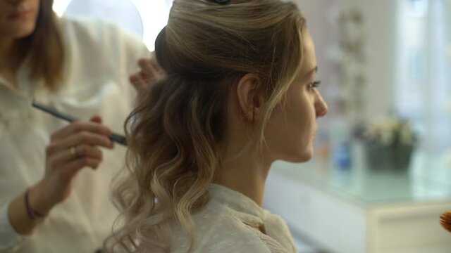 Back view of professional hairdresser making hairstyle young attractive woman and make-up artist applying makeup in beauty salon. Concept of beauty and fashion. Tracking shot in slow motion.