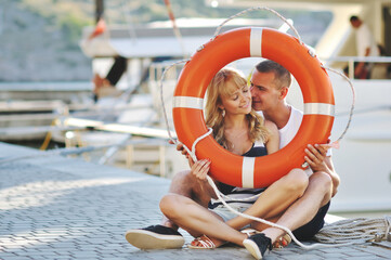 Cute young beautiful couple sitting on the ground in city near yachts with a lifebuoy and having fun together laughing and smiling kissing