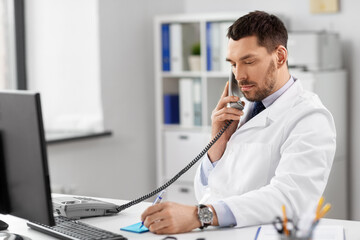Obraz premium healthcare, medicine and people concept - male doctor with clipboard calling on desk phone at hospital